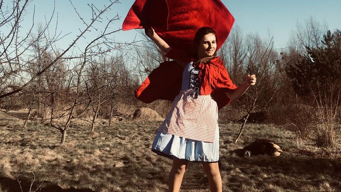 Saunamistrzyni Mariola Żurek w stroju inspirowanym Czerwonym Kapturkiem. W tle pola i zagajniki. Młoda kobieta ma na sobie czerwony kubraczek i jasną spódniczkę, w dłoni uniesionej nad głową trzyma czerwony ręcznik. 