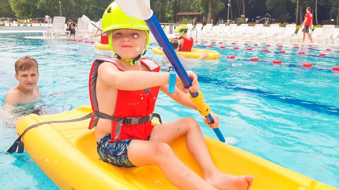 Kilkuletni uśmiechnięty chłopiec na żółtym plastikowym kajaku w zewnętrznym basenie sportowym. Ma na sobie żółty kask, czerwoną kamizelkę ratunkową, a w ręku trzyma niebiesko-białe wiosło. 