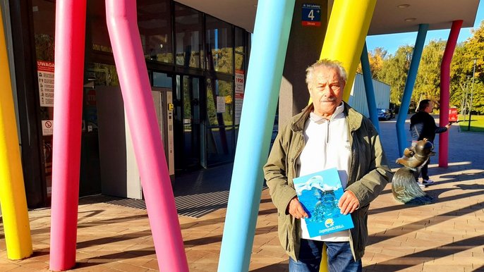 Mirosław Ścibiorski - starszy mężczyzna z wąsem pozuje na tle kolorowych filarów. Trzyma niebieską teczkę z napisem Aquapark Fala. 