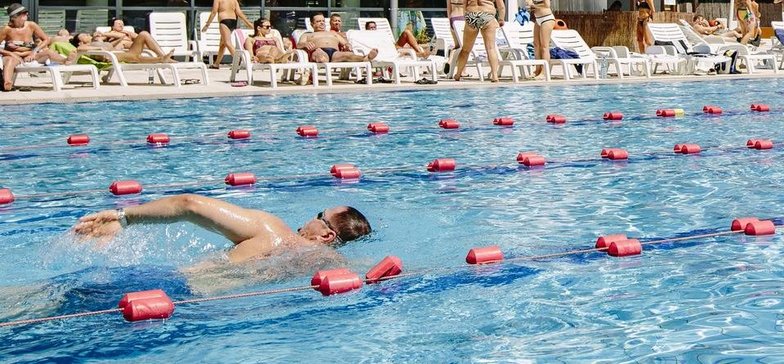 Zewnętrzny basen sportowy w słoneczny dzień. Na jednym z torów mężczyzna w niebieskich kąpielówkach.