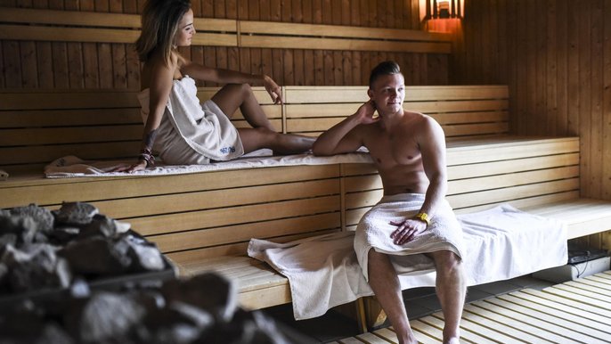 Dwoje młodych ludzi wewnątrz małej sauny zewnętrznej w Strefie Saun. Na pierwszym planie kamienie z pieca saunowego, dalej saunowicze rozłożeni na białych ręcznikach ułożonych na drewnianych ławach sauny. 
