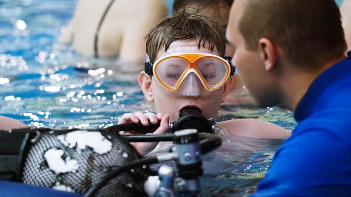 Instruktor pomaga kilkunastoletniemu chłopakowi poprawnie założyć aparat butli tlenowej. Chłopiec ma na sobie pomarańczową maskę do pływania. 