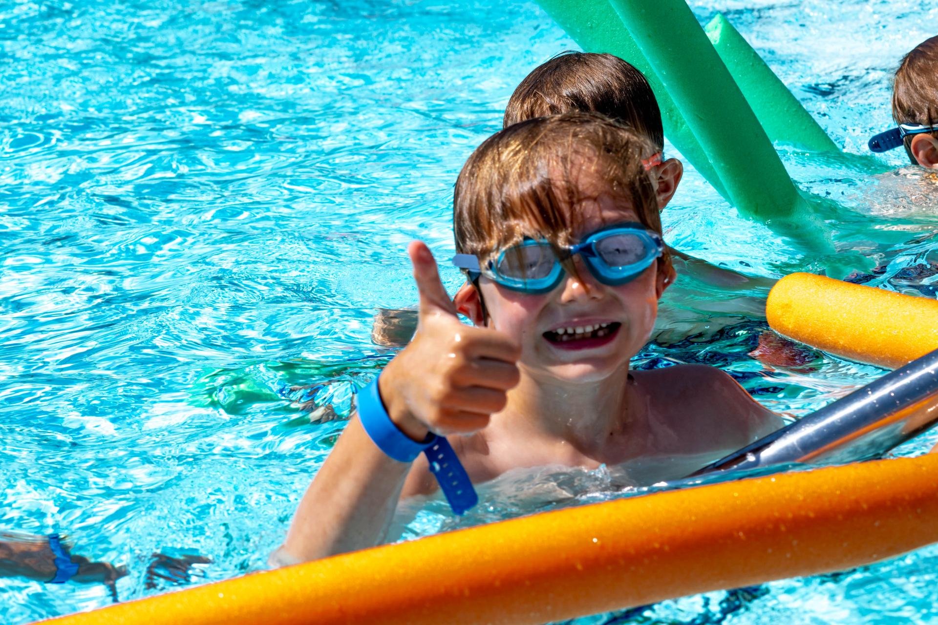 Chłopiec unoszący się na wodzie z pomocą makaronu do pływania z uśmiechem pozuje unosząc w górę kciuk. 