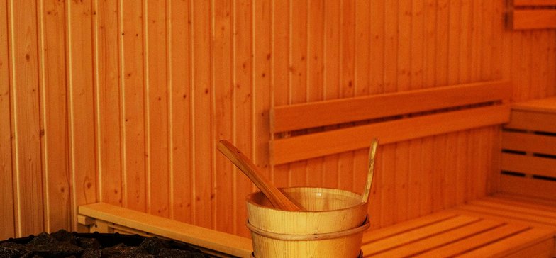 Wnętrze sauny z muzykoterapią. Drewniane wnętrze, w rogu ciemny piec otoczony drewnianą obudową, na której ułożone jest wiaderko do wody.