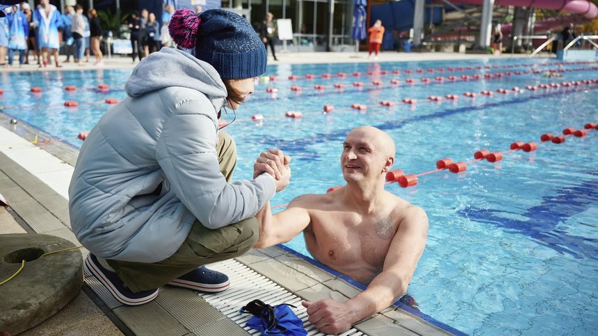 Pływak w basenie trzyma za rękę kobietę znajdującą się na brzegu basenu.