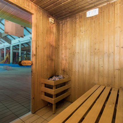 Wnętrze sauny fińskiej z perspektywy jednej z drewnianych ław, w rogu przy przeszklonych drzwiach widać piec wyłożony kamieniami, a za drzwiami można dostrzec Wodny Plac Zabaw z Muszlą. 