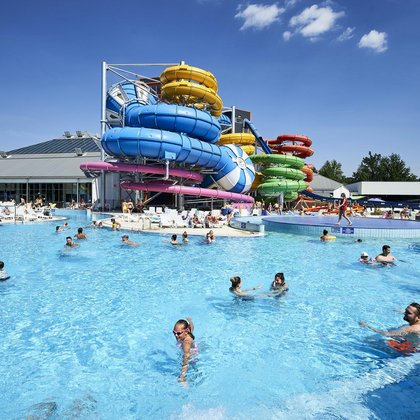 Zewnętrzny basen rekreacyjny, na pierwszym planie Falowicze kąpiący się w błękitnej wodzie. W tle kolorowe MegaZjeżdżalnie, a jeszcze dalej hala z logotypem Aquaparku FALA. 