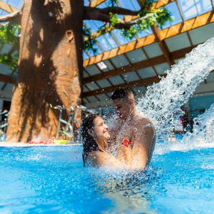 Para w wieku studenckim zażywa kąpieli w wewnętrznym basenie rekreacyjnym. Roześmianych ludzi ochlapuje woda tryskająca z jednego z masażerów karku i pleców. 
