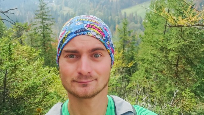 Saunamistrz Rafał Golombek - mężczyzna pozuje na tle górskiego krajobrazu. Ma na sobie zieloną koszulkę i niebieską bandanę na głowie. 