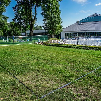 Boisko do badmintona usytuowane na trawie w części parkowej zewnętrznej Strefy Basenowej, dwa słupki i siatka. W tle widać boisko do siatkówki, a po prawej stronie zewnętrzny basen sportowy. 