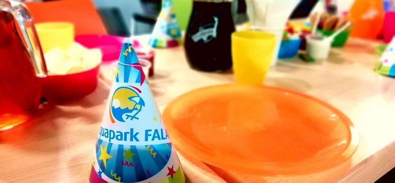 Zbliżenie: urodzinowe czapeczki z logotypem Aquaparku FALA na urodzinowym stole zastawionym kolorową zastawą i przekąskami.