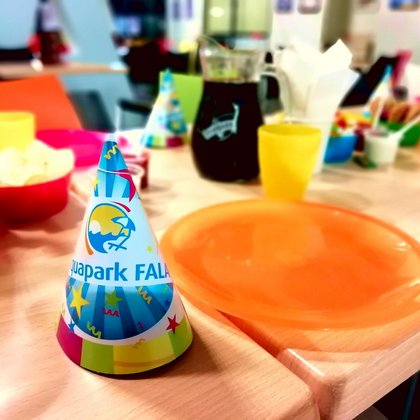 Zbliżenie: urodzinowe czapeczki z logotypem Aquaparku FALA na urodzinowym stole zastawionym kolorową zastawą i przekąskami. 