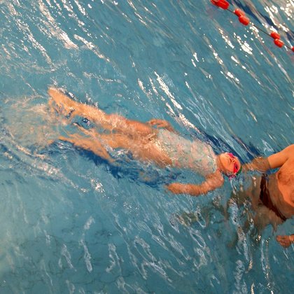 Instruktor stoi w basenie sportowym, przed sobą ma kilkuletnią dziewczynkę w błękitnym stroju kąpielowym, unoszącą się na wodzie na plecach. 