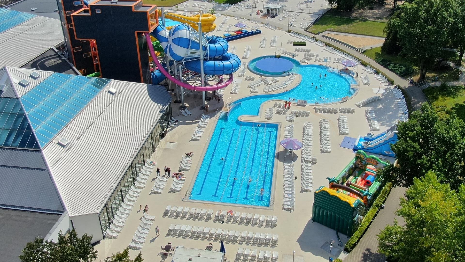 Panorama terenu zewnętrznego z perspektywy drona. Na pierwszym planie basen sportowy z czterema torami, dalej widać kolorowe zjeżdżalnie i basen rekreacyjny z rwącą rzeką. 