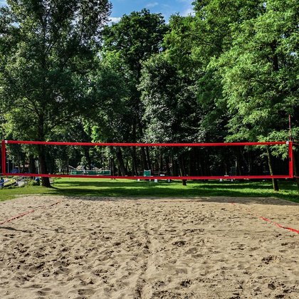 Boisko do siatkówki plażowej w słoneczny dzień. W tle część parkowa zewnętrznej Strefy Basenowej, wysokie, zielone drzewa.