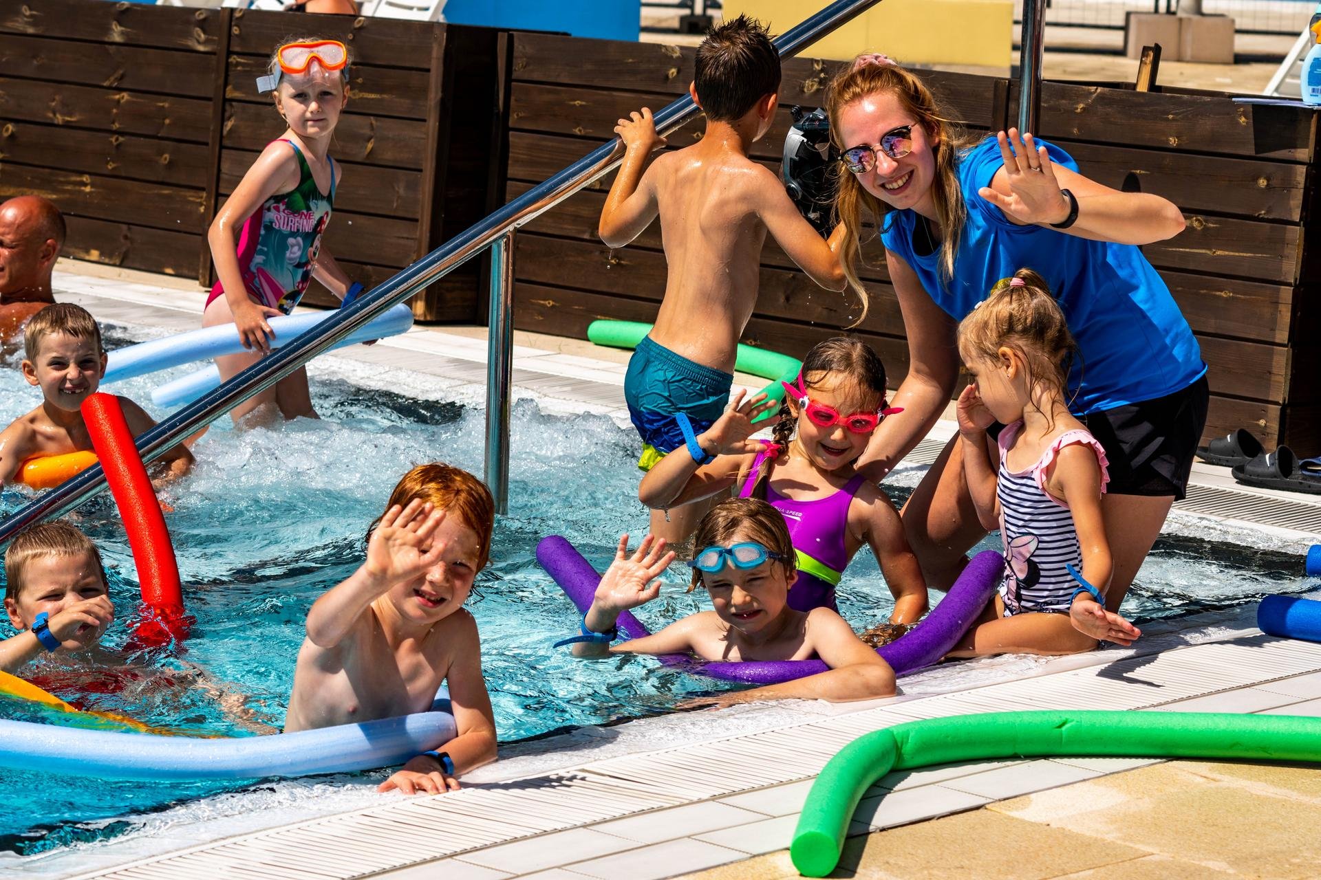 Grupa dzieci wraz z instruktorem przy brzegu basenu wypływowego w słoneczny dzień. Z uśmiechem patrzą w obiektyw i machają radośnie. 