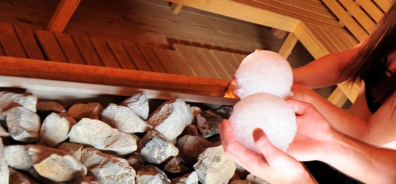 Zbliżenie na piece w dużej saunie zewnętrznej. Widać dłonie dwóch saunamistrzów, w których trzymają oni lodowe kule.