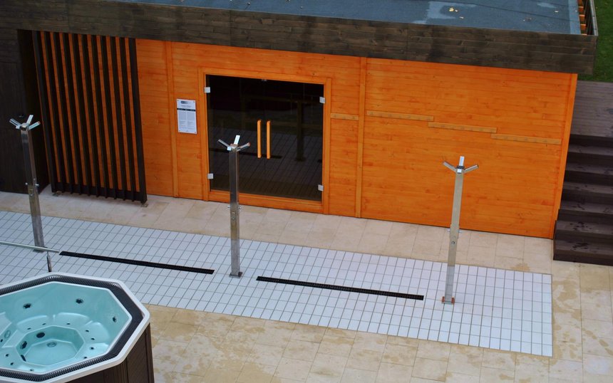 Duża sauna zewnętrzna z zewnątrz. Zabudowana drewnem w kolorze pomarańczowym, z brązowymi elementami dekoracyjnymi. Przed wejściem do sauny trzy poczwórne prysznice zewnętrzne i jacuzzi.