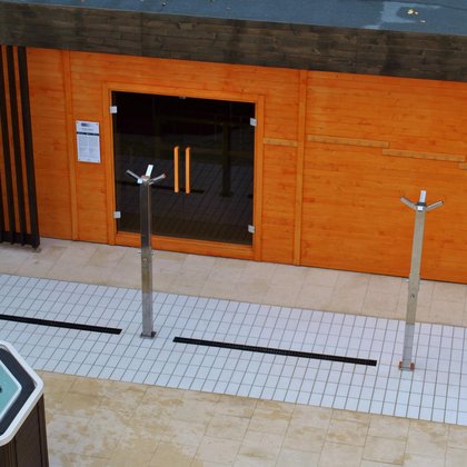 Duża sauna zewnętrzna z zewnątrz. Zabudowana drewnem w kolorze pomarańczowym, z brązowymi elementami dekoracyjnymi. Przed wejściem do sauny trzy poczwórne prysznice zewnętrzne i jacuzzi. 