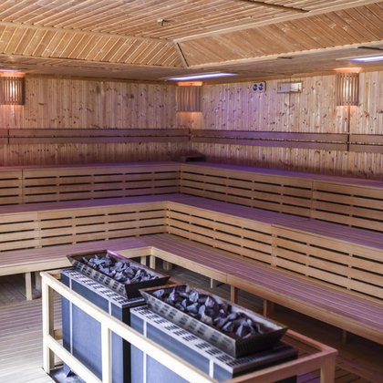 Wnętrze dużej sauny zewnętrznej. Obudowana całkowicie drewnem, w centralnym punkcie podwójny piec saunowy z ciemnymi kamieniami. Wzdłuż ścian trzy rzędy drewnianych ław. 