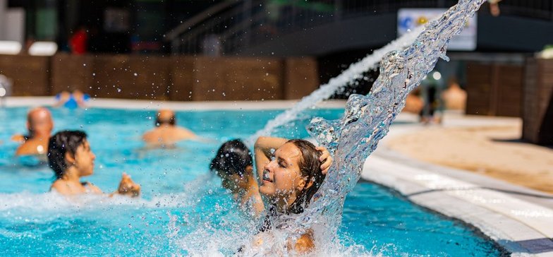 Zdjęcie młodej kobiety, która jest w basenie. Kobieta jest uśmiechnięta. Stoi pod dyszą z wodą. 