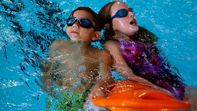 Dwoje dzieci w basenie sportowym. Płyną na plecach trzymając się razem pomarańczowej bojki ratowniczej. Chłopiec po lewej nosi granatowe okularki pływackie, dziewczynka po prawej fioletowy kostium i niebieskie okularki. 