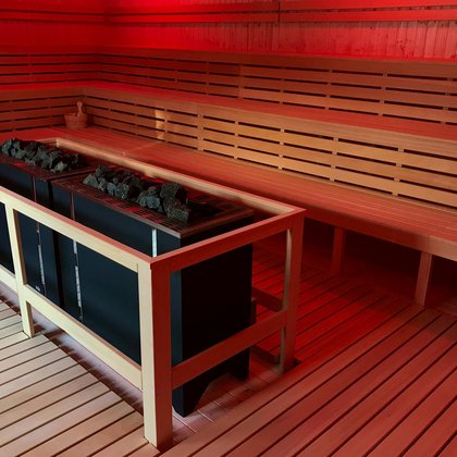 Wnętrze dużej sauny fińskiej zewnętrznej. Zbliżenie na dwa piece w centralnym punkcie sauny. Dookoła drewniane ławy z trzema rzędami siedzisk. Czerwone oświetlenie. 