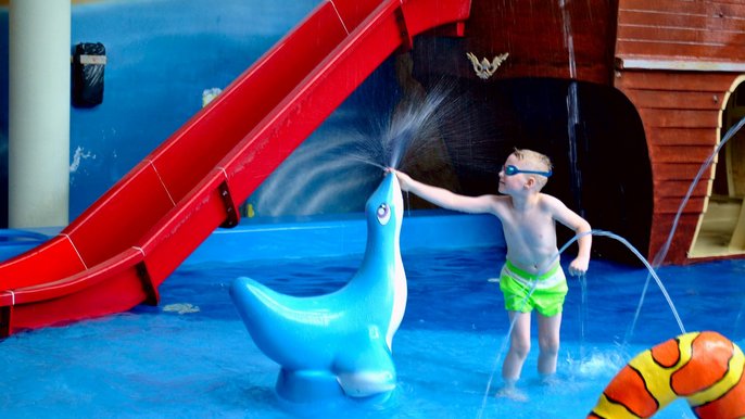 Wodny Plac Zabaw ze Statkiem. Kilkuletni chłopiec w zielonych kąpielówkach i niebieskich okularach pływackich bawi się przy fontannie-foce. 