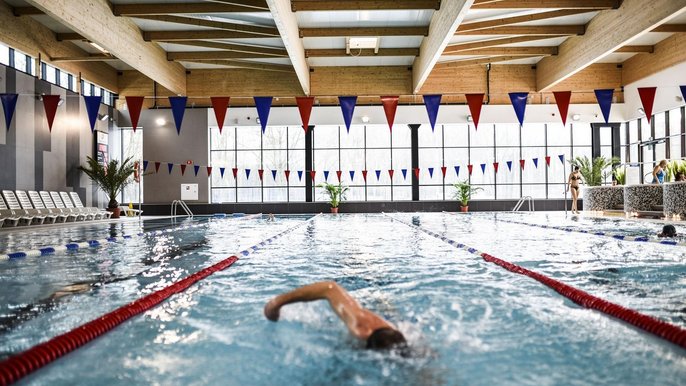 Basen sportowy wewnętrzny. Na środkowym torze pływak unosi rękę nad wodą, resztę ciała znajduje się pod powierzchnią. Po prawo widać wanny jacuzzi, a w tle przeszkloną ścianę hali basenowej. 