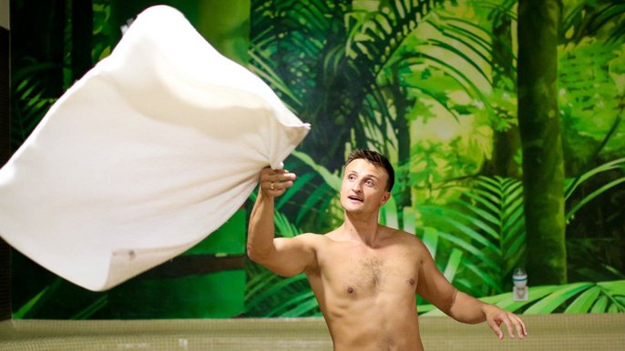Saunamistrz Radosław Sieraj - młody mężczyzna w ręczniku wokół bioder lewą ręką trzymający ręcznik rozłożony w locie ręcznik. W tle ciemna ściana z zielonymi wzorami dżungli. 