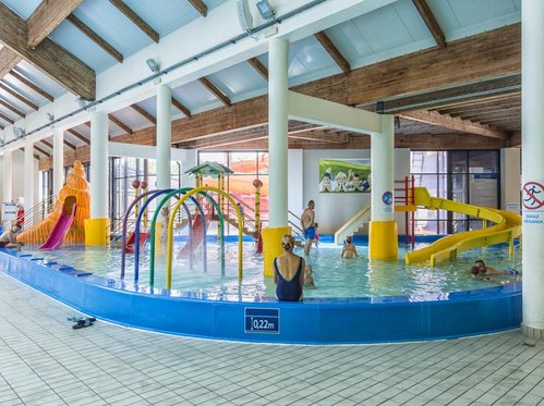 Panorama wodnego placu zabaw z muszlą. Na pierwszym planie mama nadzorująca zabawę dzieci, kolorowe kurtyny wodne, w tle dom na palach. Po prawo żółta zjeżdżalnia, a po lewo zjeżdżalnia-muszla.