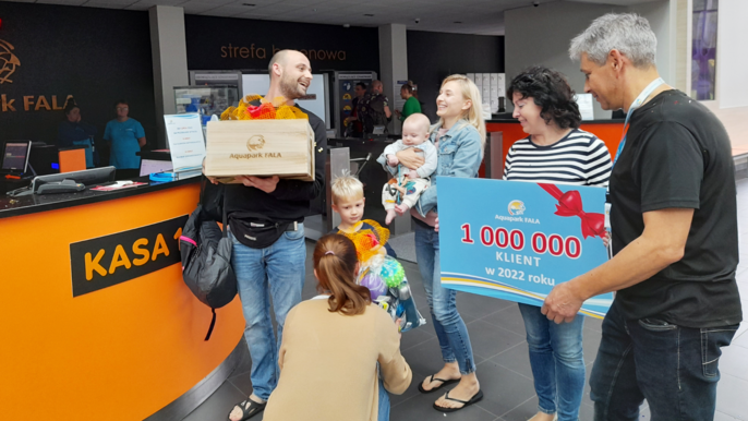 Na zdjęciu jest 7 osób: 3 kobiety, 2 mężczyzn i 2 dzieci w tym niemowlę trzymane na rękach przez młodą uśmiechniętą blondynkę. Mężczyzna po prawej trzyma tabliczkę z napisem 1000000 klient w 2022 r. Osoby stoją obok kas Aquaparku FALA. 
