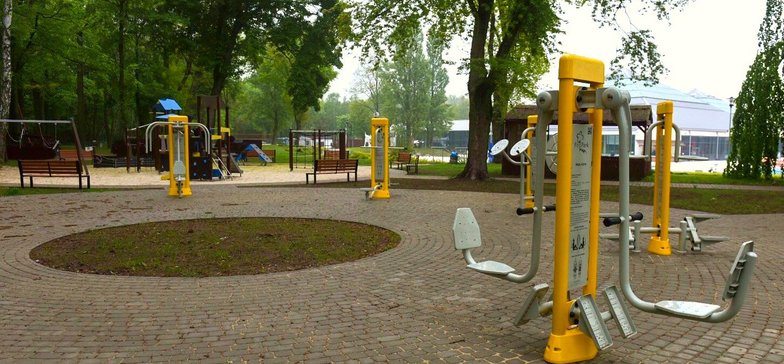 Siłownia zewnętrzna usytuowana w części parkowego Aquaparku FALA. Na szarej kostce brukowej stoją żółto-szare sprzęty. W tle plac zabaw dla dzieci i jedna z hal-piramid Aquaparku.