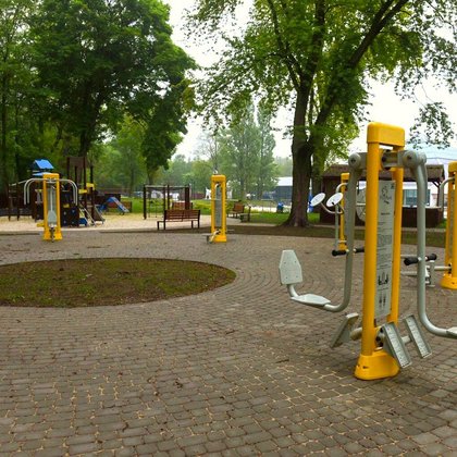 Siłownia zewnętrzna usytuowana w części parkowego Aquaparku FALA. Na szarej kostce brukowej stoją żółto-szare sprzęty. W tle plac zabaw dla dzieci i jedna z hal-piramid Aquaparku. 