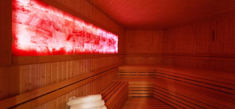 Wnętrze sauny solnej całkowicie zabudowane drewnem. Po lewej stronie na ścianie jest prostokątny panel solny podświetlony w kolorze czerwonym, pod nim jedna ława, wzdłuż pozostałych dwóch ścian dwa rzędy drewnianych siedzisk.