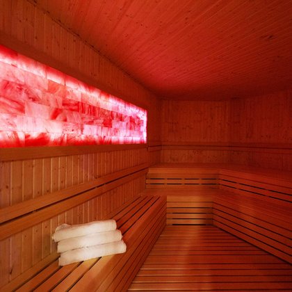 Wnętrze sauny solnej całkowicie zabudowane drewnem. Po lewej stronie na ścianie jest prostokątny panel solny podświetlony w kolorze czerwonym, pod nim jedna ława, wzdłuż pozostałych dwóch ścian dwa rzędy drewnianych siedzisk. 