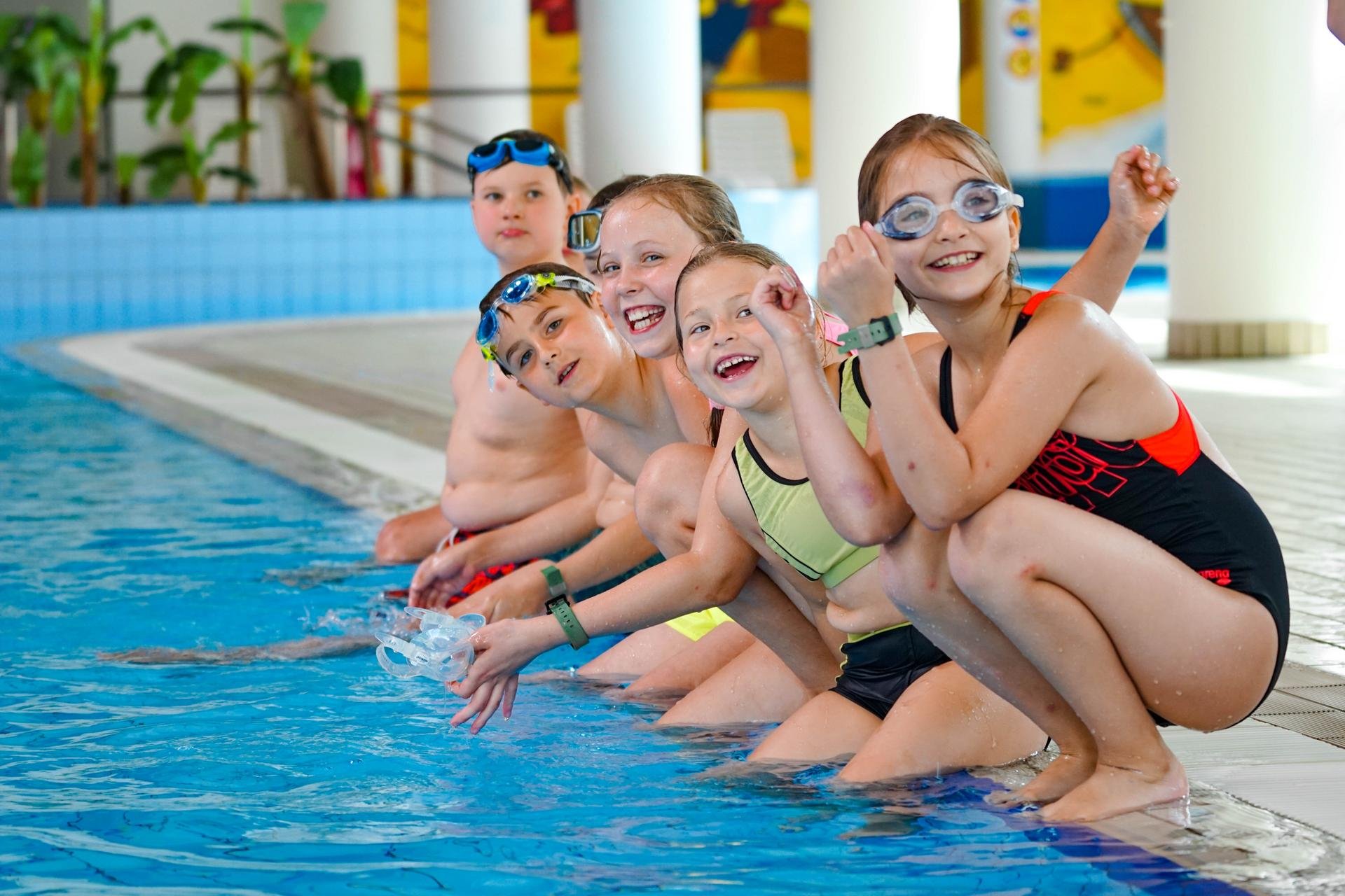 Kilkoro dzieci siedzi i kuca nad brzegiem basenu z uśmiechem spoglądając w obiektyw. Mają na sobie kolorowe stroje do pływania i okularki pływackie. 