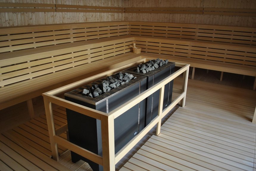 Wnętrze dużej sauny zewnętrznej. W centralnej części ciemny piec saunowy, dookoła trzy rzędy drewnianych ław, również podłoga sauny wyłożona jest drewnem.