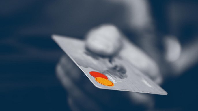Zbliżenie na dłoń trzymającą plastikową kartę - zdjęcie w odcieniach szarości, jedynie logo na karcie w intensywnie czerwonych i pomarańczowych barwach. 