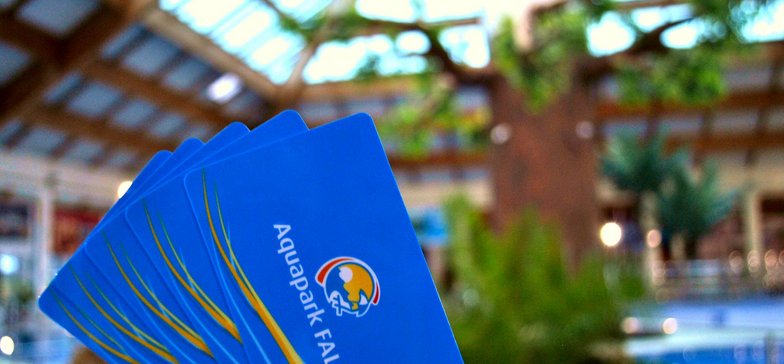 Plik niebieskich kart z logotypem Aquaparku FALA. W tle drzewo tropikalne przy wewnętrznym basenie rekreacyjnym.