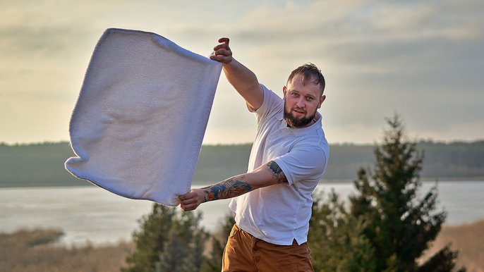 Saunamistrz Tomasz Jabłoński, mężczyzna w białej koszulce i brązowych spodniach, unoszący w rękach biały ręcznik. Pozuje na tle jeziora i drzew. 