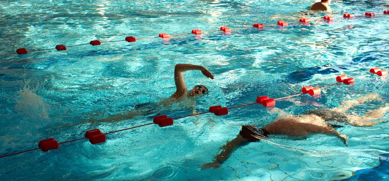 Wewnętrzny basen sportowy. W turkusowej wodzie na wyznaczonych torach stylem klasycznym pływa kilkoro ludzi.