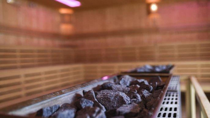 Zbliżenie na piec saunowy w dużej saunie zewnętrznej Strefy Saun. Ciemne kamienie na pierwszym planie, w tle dwie ściany sauny obudowane drewnem wraz z trzema rzędami siedzisk. Lekkie oświetlenie o różowej barwie. 