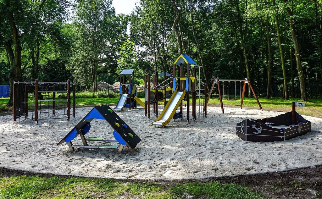 Suchy plac zabaw wybudowany na piaszczystym okręgu. Zjeżdżalnie, elementy do wspinaczki, huśtawki i piaskownica są drewniane inspirowane stylem pirackim. W tle strefa parkowa.