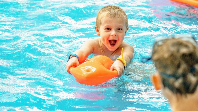 Mały chłopiec o blond włosach i z szerokim uśmiechem w basenie trzyma bojkę ratowniczą i spogląda na kolegów czekających na brzegu. 