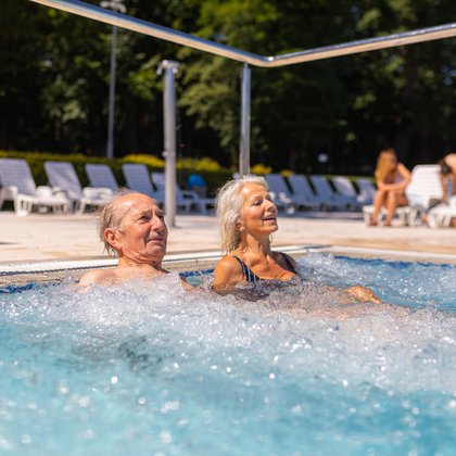 Zewnętrzny basen rekreacyjny w słoneczny dzień. Para seniorów korzysta z leżaków do masażu powietrznego umieszczonych przy brzegu. W tle część parkowa Aquaparku. 