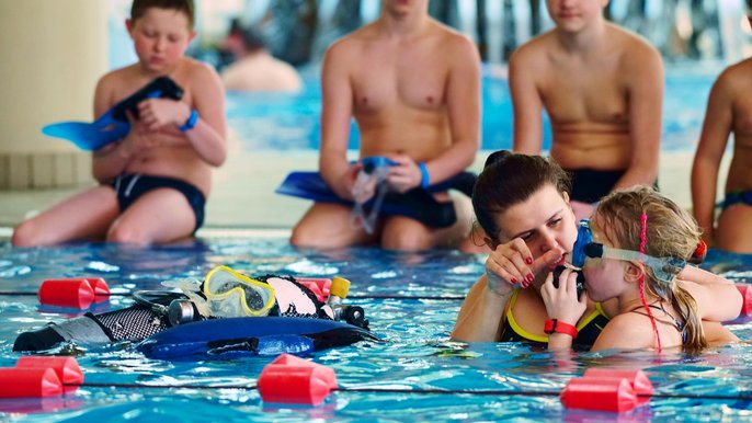 Instruktorka pomaga małej dziewczynce korzystać z aparatu tlenowego podłączonego do butli do snurkowania. Na brzegu basenu czekają na swoją kolej kolejne dzieci. 