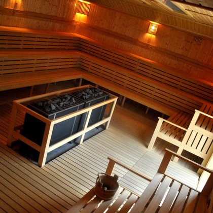 Panorama wnętrza dużej sauny zewnętrznej. W centralnym punkcie podwójny, prostokątny piec saunowy pokryty ciemnymi kamieniami. Sauna całkowicie zabudowana drewnem - wzdłuż wszystkich ścian trzy rzędy siedzisk. 