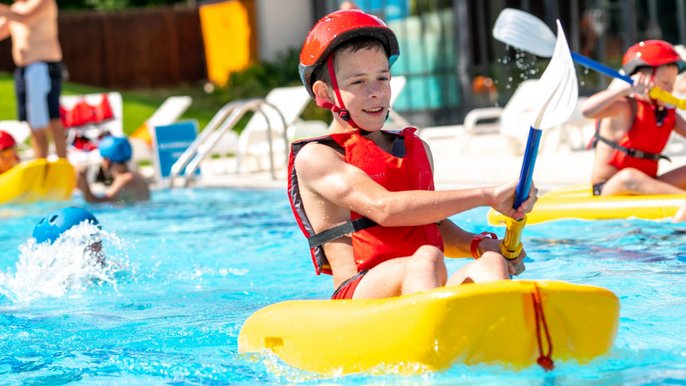 Kilkunastoletni chłopak w żółtym kajaku i czerwonym kapoku na jednym z torów basenu sportowego. 