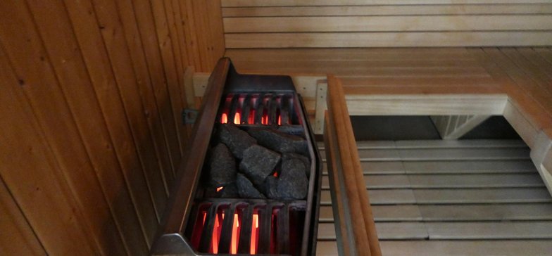 Wnętrze wewnętrznej sauny fińskiej. Dookoła drewniane ławy, po lewo piec saunowy wyłożony kamieniami.